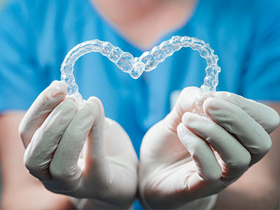 The image shows a pair of hands holding a heart-shaped transparent object, which appears to be a dental retainer or mouthguard, with one hand wearing gloves.