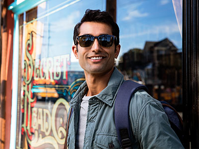 A man wearing sunglasses stands outside a storefront, smiling slightly, with his arms crossed, holding a backpack, and looking towards the camera.