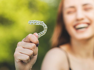 The image shows a person holding up a toothbrush with a smile, set against a blurred outdoor background.