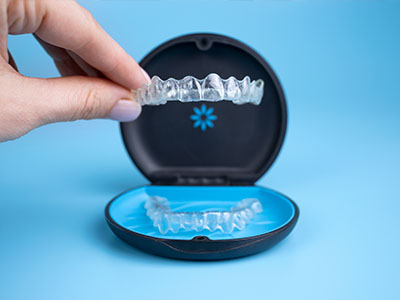 An open box containing a set of clear, translucent plastic aligners, possibly for dental use, with a human hand holding up one of the aligners.