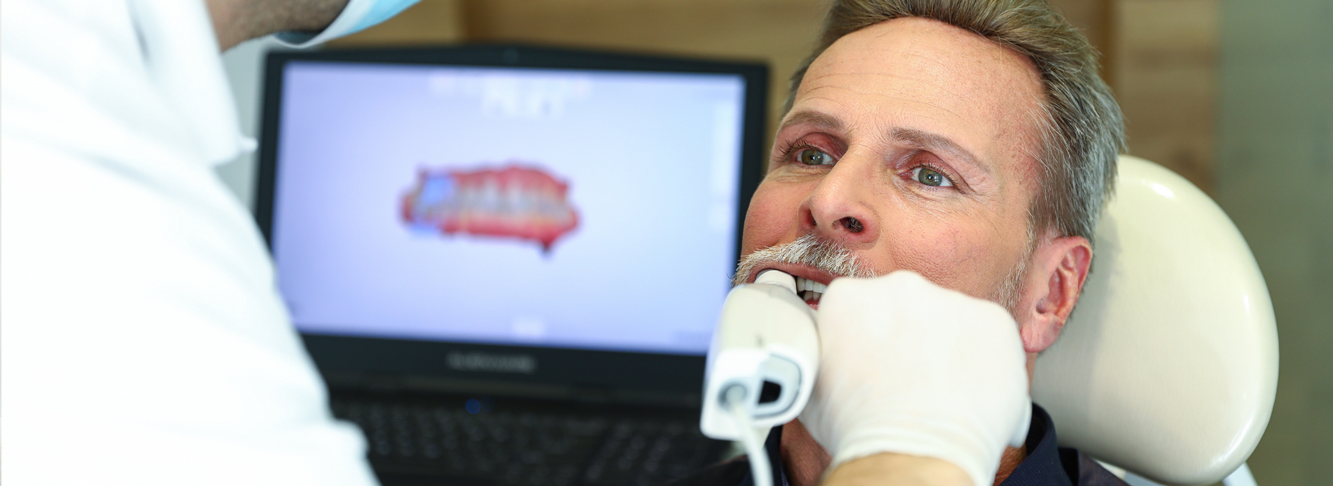 A man is sitting in a dental chair, receiving dental care from a dentist who is standing beside him.