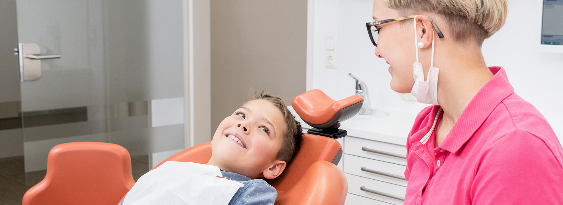 The image shows a young boy sitting in a dental chair with a big smile on his face, receiving dental care from a woman who appears to be a dentist, in an office setting.