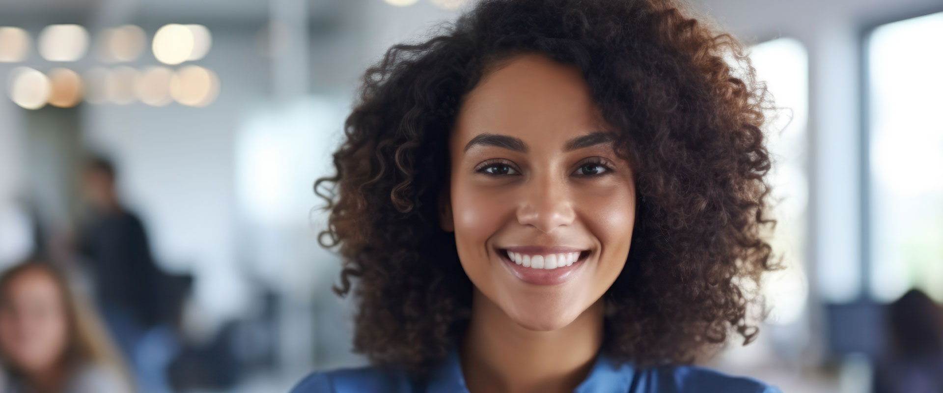 A smiling woman with curly hair, wearing a blue shirt, stands confidently in an office environment.