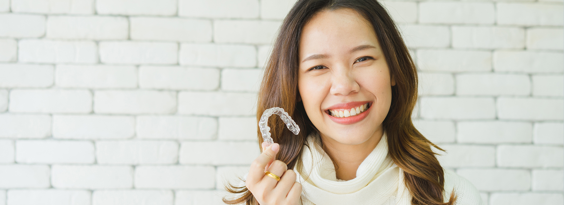The image shows a smiling woman holding a toothbrush up to her mouth with a brick wall background and a white banner across the top of the photo.