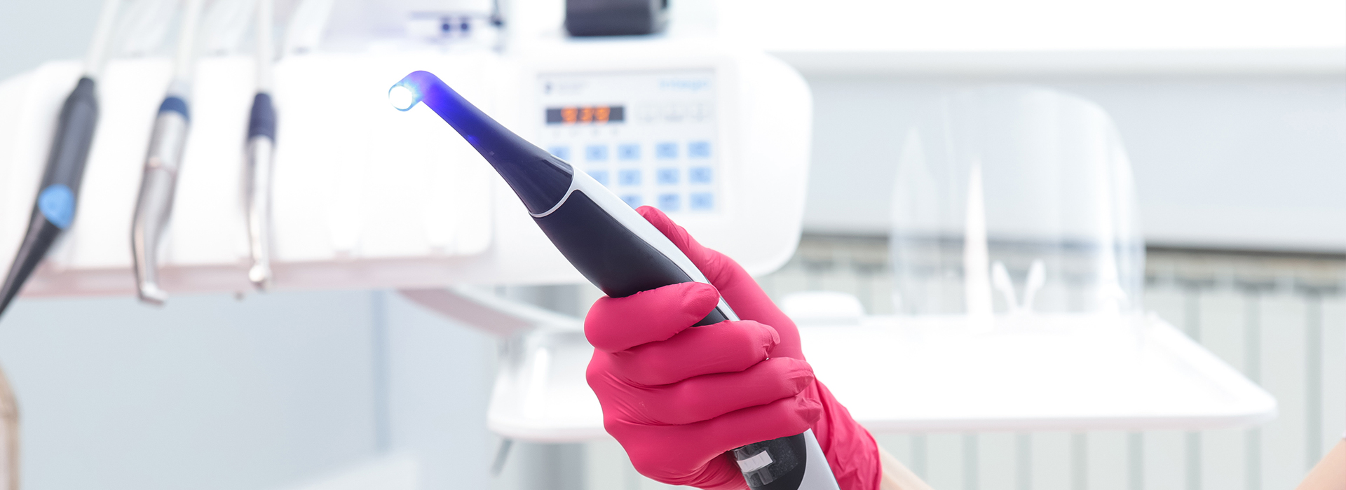 The image shows a person wearing a red glove holding an electronic device with a blue light, possibly a stylus, near a white object that resembles a microphone or a similar electronic component, in a setting that appears to be a laboratory or a workshop.