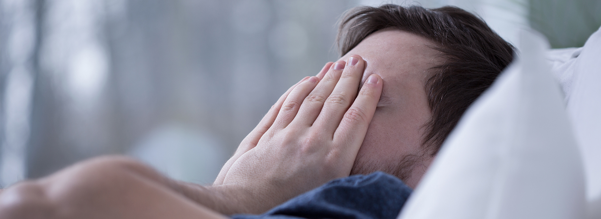 The image shows a man lying on a bed with his hands covering his face, appearing to be asleep or resting.