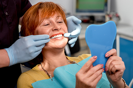 In the image, a woman is seated with her teeth being cleaned by a dental professional. She is holding up a blue dental model and smiling at the camera while wearing a face mask.
