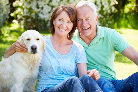 A man and woman sitting outdoors with a golden retriever between them, smiling at the camera.