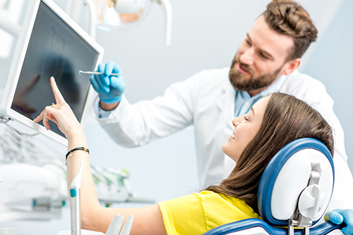 A man in a white coat standing next to a woman seated in a dental chair with a screen behind them, both are in a modern dental office setting.