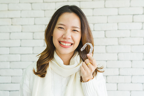 A woman holding a smiley face emoji with a content expression in front of a brick wall.