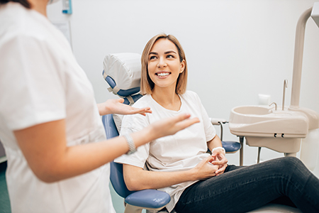The image shows a woman seated in a dental chair, smiling at the camera, with a dental professional standing behind her, both in a dental office setting.