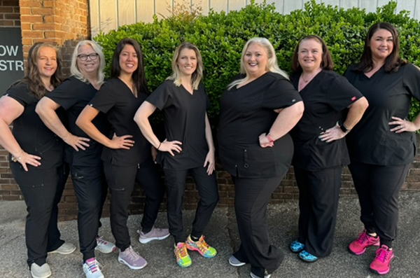 A group of women wearing scrubs stand together for a photo, posing confidently in front of a brick building.