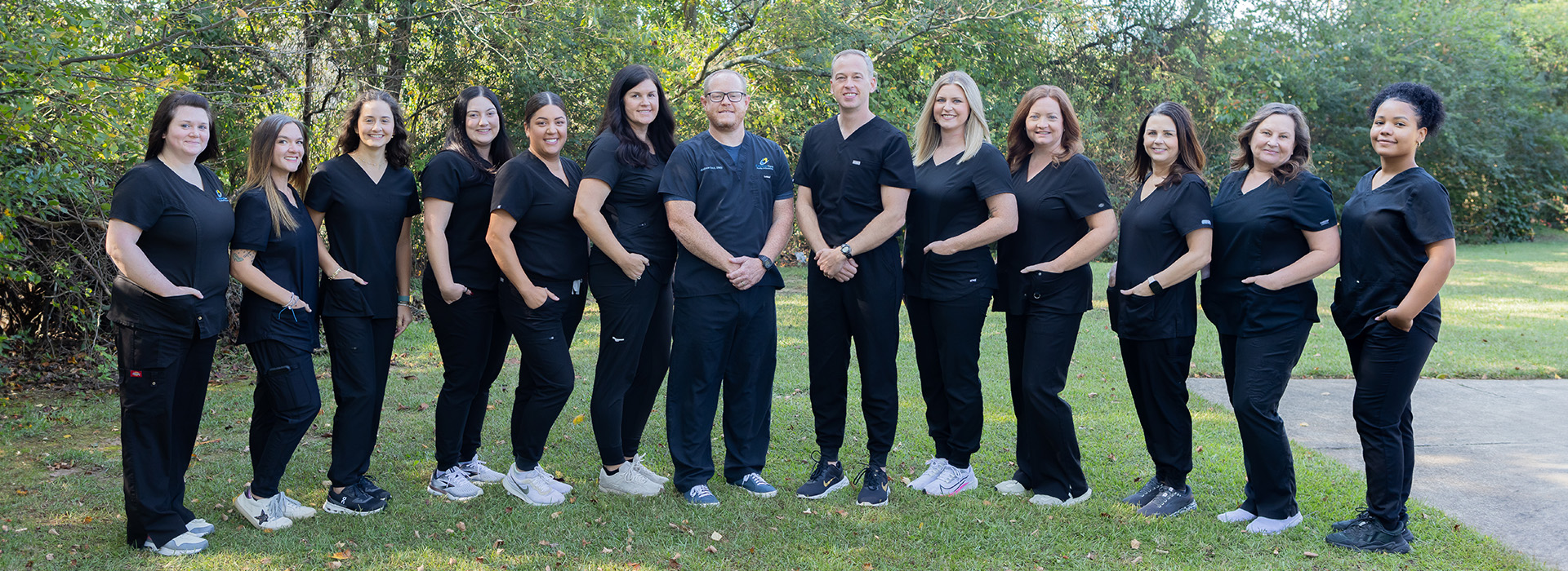 The image shows a group of individuals, likely professionals, posing together in front of a building with a sign that reads DENTAL CLINIC . They appear to be dressed in scrubs and are smiling at the camera.
