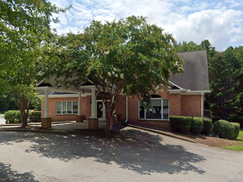 The image depicts a two-story building with a prominent front entrance, featuring a white columned portico and a covered entryway. The structure is topped by a gabled roof with asphalt shingles, and the facade is adorned with a mix of brickwork and siding. A large tree with green leaves stands in front of the building, partially obscuring the view. The sky above is partly cloudy, suggesting a fair weather day.