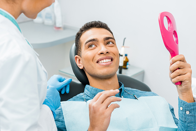 A young man sitting in a dental chair with an open mouth, holding a pink object and smiling, while being attended to by a dentist wearing a blue apron.